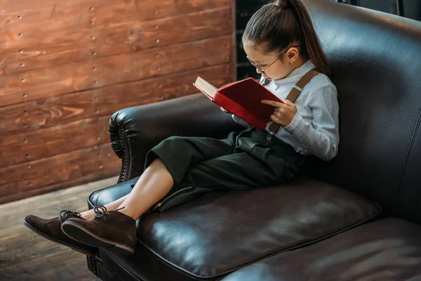 Stylish little child reading book on couch at home — Stock Photo