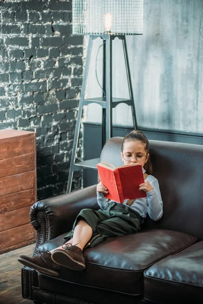Niño pequeño concentrado leyendo el libro en el sofá en casa - foto de stock