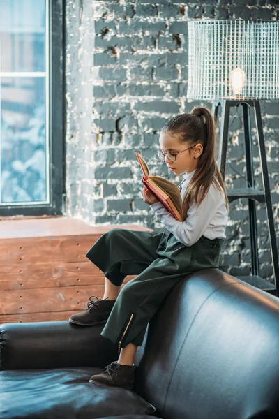 Konzentriertes kleines Kind liest Buch, während es auf der Couch in Loft-Wohnungen sitzt — Stockfoto