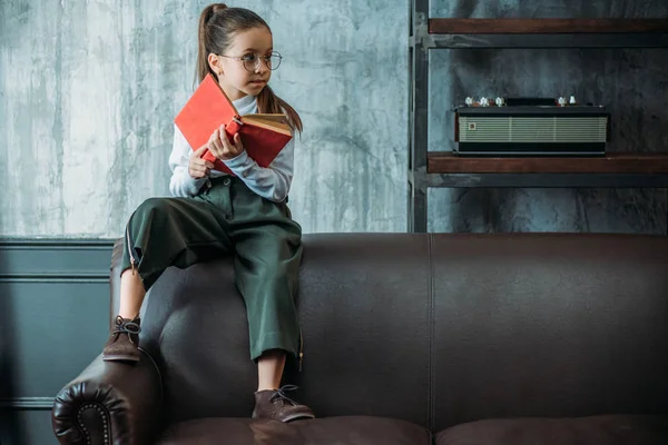 Petit enfant réfléchi avec livre assis sur le canapé dans des appartements loft — Photo de stock