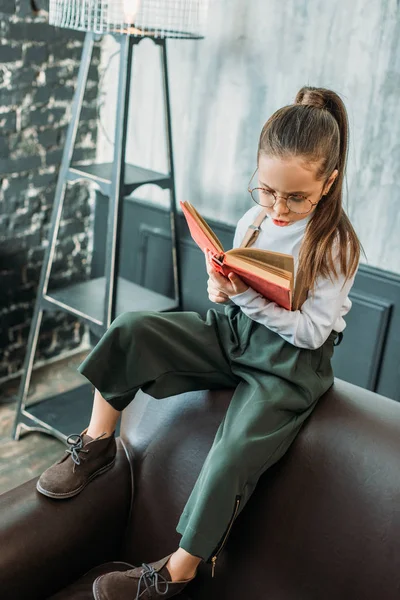 Зосереджена маленька дитина читає книгу, сидячи на дивані — стокове фото