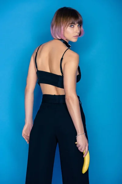 Rear view of fashionable girl holding banana and looking at camera isolated on blue — Stock Photo