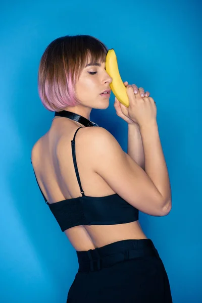 Chica con estilo en la parte superior de la cosecha tocando la cara con plátano aislado en azul - foto de stock