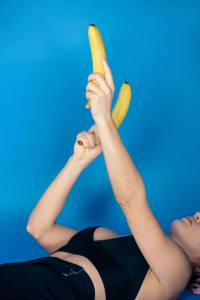 Imagem cortada de menina elegante deitado e segurando bananas em mãos na frente dela em azul — Fotografia de Stock