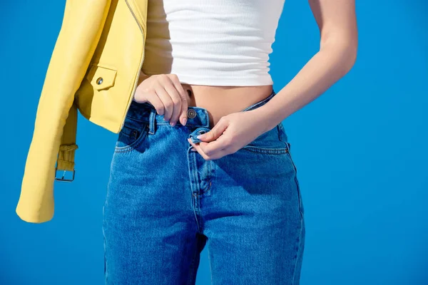 Обрізане зображення модної дівчини, що розстібає джинси, ізольовані на синьому — стокове фото