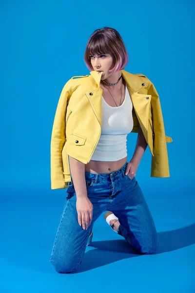 Fashionable girl sitting on knees, biting leather jacket and looking away on blue — Stock Photo