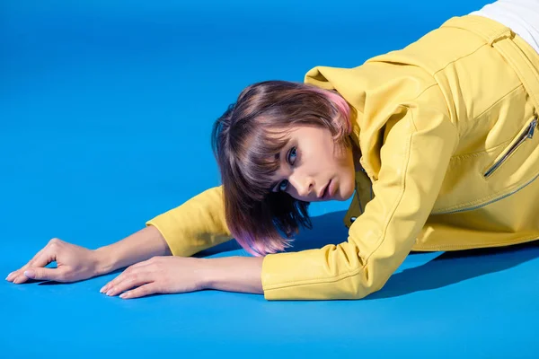 Seductive stylish girl lying in yellow leather jacket and looking at camera on blue — Stock Photo