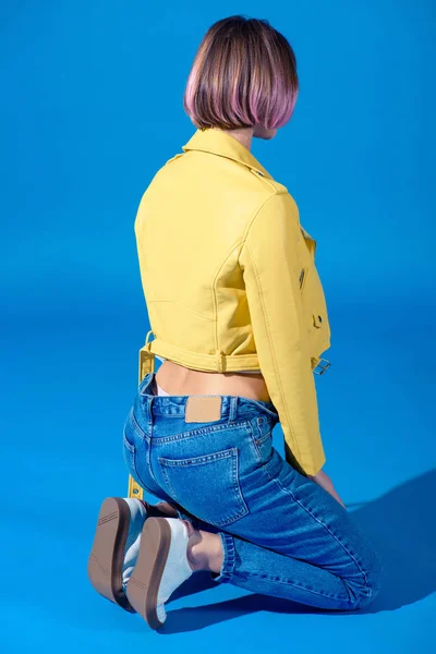 Rear view of stylish girl sitting on knees in jacket and jeans on blue — Stock Photo