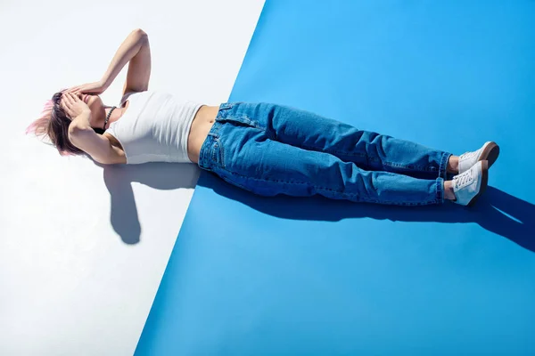 Menina na moda deitada no chão branco e azul em jeans e cobrindo os olhos com as mãos — Fotografia de Stock