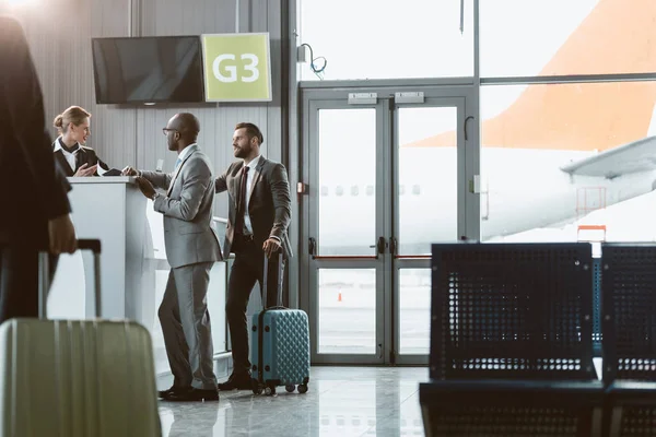 Hombres de negocios de pie en la recepción del aeropuerto para comprar boletos mientras su colega camina hacia ellos - foto de stock