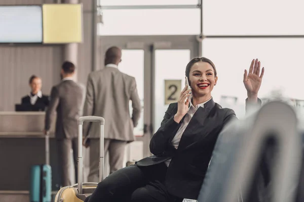 Junge Geschäftsfrau wartet in der Flughafenlobby auf ihr Flugzeug, während sie telefoniert und winkt — Stockfoto