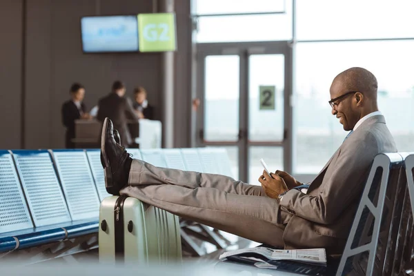 Homme d'affaires souriant utilisant un smartphone en attendant son vol dans le hall de l'aéroport — Photo de stock