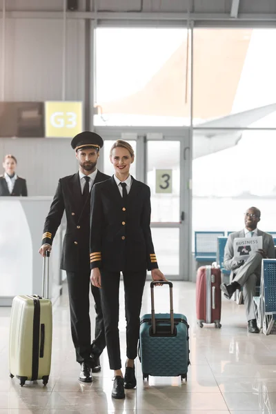 Молодые пилоты мужского и женского пола прогуливаются по вестибюлю аэропорта с чемоданами — стоковое фото