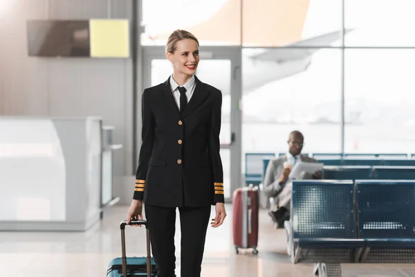 Pilota donna sorridente con valigia che cammina presso la hall dell'aeroporto — Foto stock