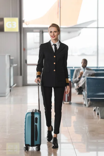 Щаслива жінка-пілот з валізами, що йдуть у вестибюлі аеропорту — стокове фото