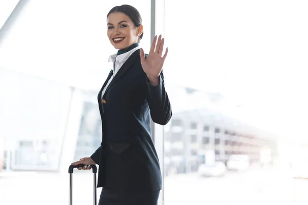 Atractiva azafata joven con maleta saludando a la cámara en el aeropuerto - foto de stock