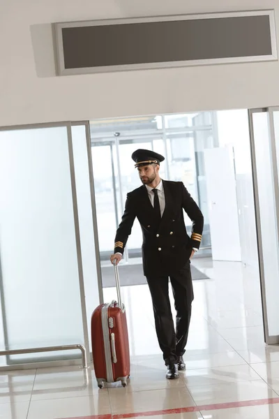 Красивый молодой пилот с чемоданом в зоне вылета аэропорта — стоковое фото