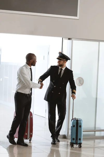 Guapo piloto apretón de manos con joven hombre de negocios en la zona de salida del aeropuerto - foto de stock