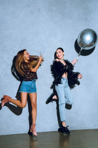 Estilo africano americano chica con smartphone fotografiando asiático amigo divertirse con globo en fiesta - foto de stock