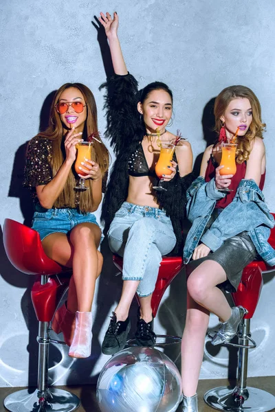 Schöne modische multiethnische Frauen sitzen auf Barhockern und trinken Cocktails auf einer Party — Stockfoto
