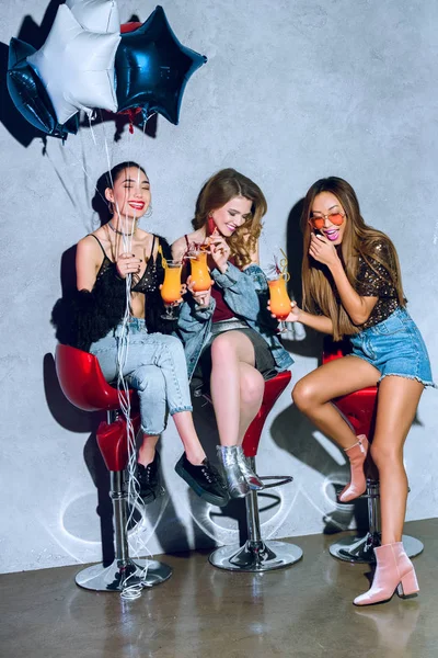 Hermosas mujeres multiétnicas con estilo con globos bebiendo cócteles en la fiesta - foto de stock
