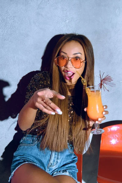 Surprised fashionable mulatto girl holding cocktail and pointing at camera — Stock Photo