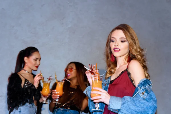 Beautiful smiling young stylish woman holding glass with cocktail while multiethnic friends standing behind — Stock Photo