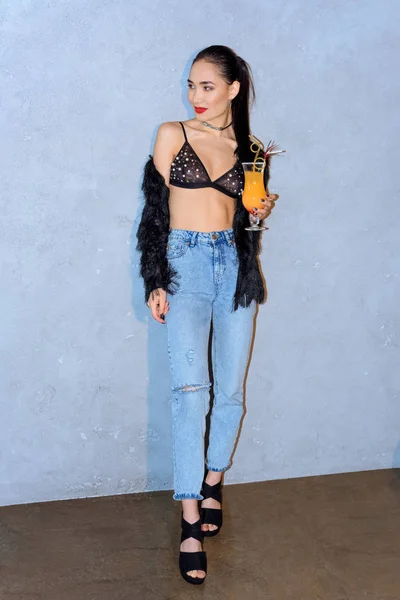 Модная молодая азиатская женщина, держа стакан коктейля и глядя в сторону серого — стоковое фото
