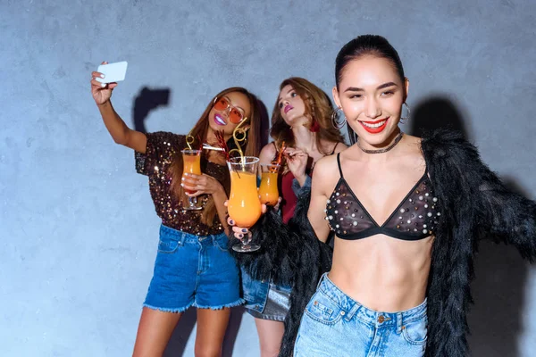 Lächelnd stilvolle asiatische Frau hält Glas mit Cocktail, während Freunde Cocktails trinken und Selfie mit Smartphone auf der Party machen — Stockfoto