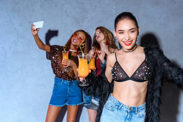 Souriant élégant asiatique femme tenant verre avec cocktail tandis que les amis boire des cocktails et de prendre selfie avec smartphone derrière à la fête — Photo de stock