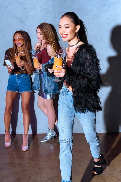 Sorridente donna asiatica elegante che tiene il bicchiere con cocktail mentre gli amici bevono cocktail e utilizzano lo smartphone dietro alla festa — Foto stock