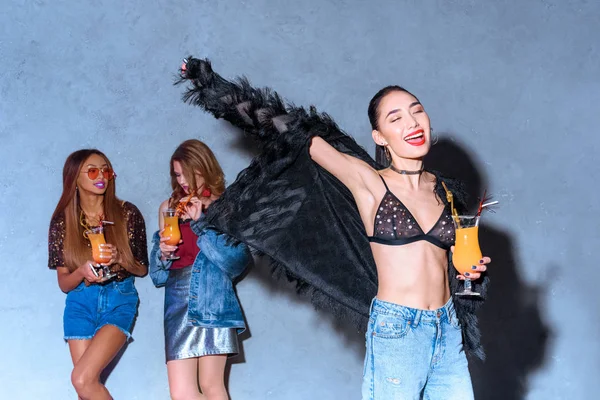 Cheerful stylish asian girl holding glass with cocktail while friends drinking behind at party — Stock Photo