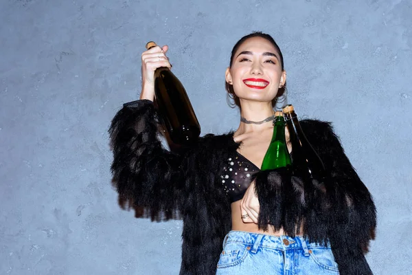 Fashionable happy asian girl holding bottles with alcoholic beverages on grey — Stock Photo