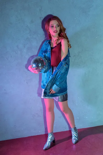 Mujer joven de moda sosteniendo la bola disco y mirando hacia otro lado en gris - foto de stock
