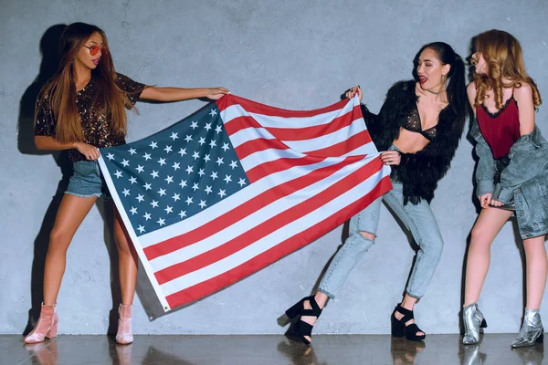 Mujeres jóvenes multiculturales con bandera americana contra muro de hormigón — Stock Photo