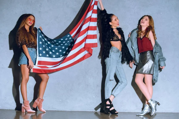 Mujeres jóvenes multiculturales con bandera americana contra muro de hormigón - foto de stock