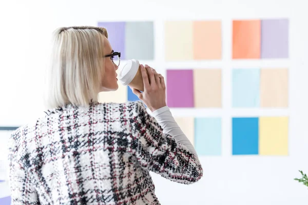 Attractive fashion magazine editor drinking coffee and looking at color palette on wall — Stock Photo