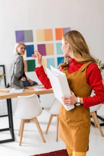 Attractive magazine editor with folder pointing at colleague and color palette on wall behind — Stock Photo