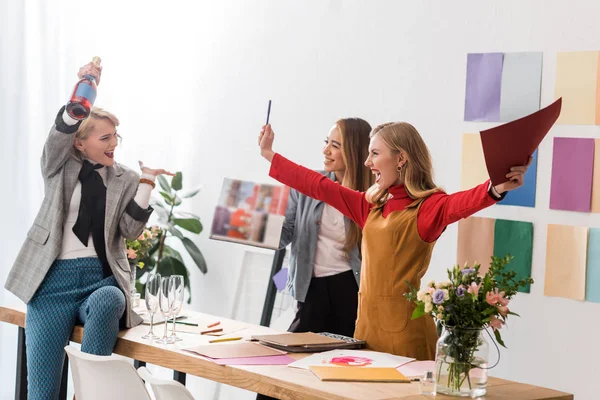 Emocionados editores de revistas gritando y celebrando con champán en la oficina moderna - foto de stock