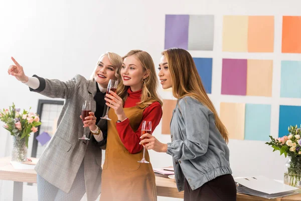 Editores de revistas multiétnicas celebrando con champán y señalando en algún lugar de la oficina moderna - foto de stock