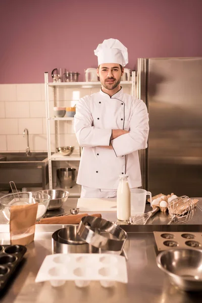 Joven pastelero con los brazos cruzados de pie en el mostrador en la cocina del restaurante - foto de stock