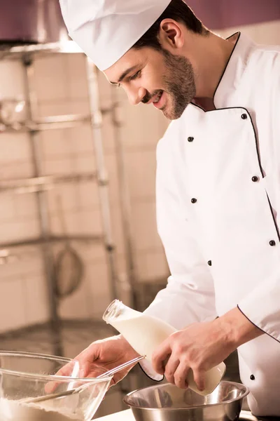 Pasticceria in cappello da chef con latte in mano facendo impasto in cucina ristorante — Foto stock