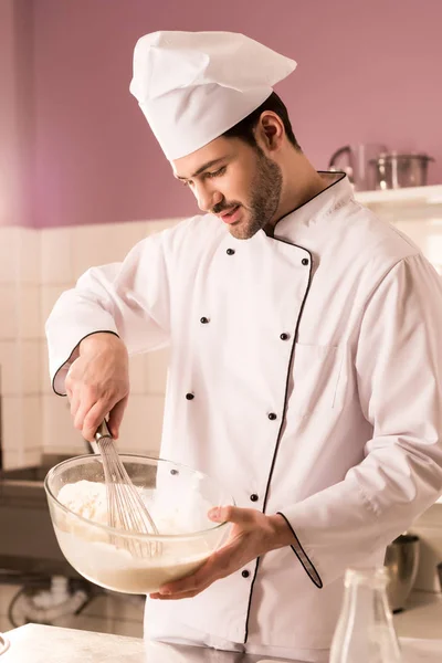 Pastelero joven en sombrero de chef haciendo masa en la cocina del restaurante - foto de stock