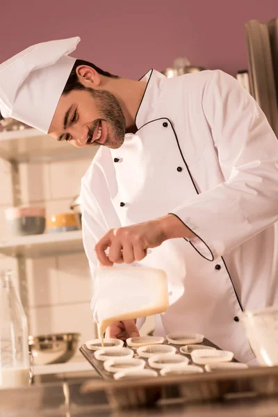 Улыбающийся кондитер наливает тесто в хлебопекарные формы на кухне ресторана — стоковое фото