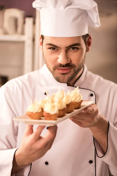 Retrato de joven pastelero mirando cupcakes en plato en las manos - foto de stock