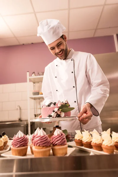 Confitero sonriente de pie en el mostrador con pastel y cupcakes en la cocina del restaurante — Stock Photo