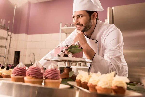 Confiseur souriant debout au comptoir avec gâteau et cupcakes dans la cuisine du restaurant — Photo de stock