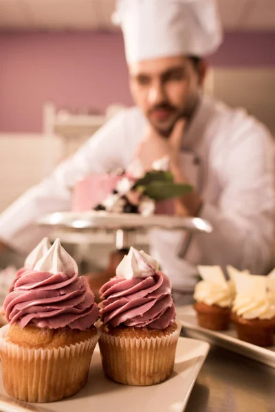 Foco seletivo de cupcakes e confeiteiro de pé perto do bolo no balcão na cozinha do restaurante — Fotografia de Stock
