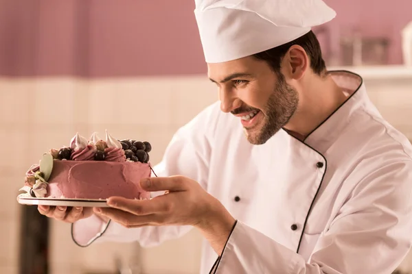Vista lateral del pastelero sonriente mirando el pastel en las manos en la cocina del restaurante - foto de stock