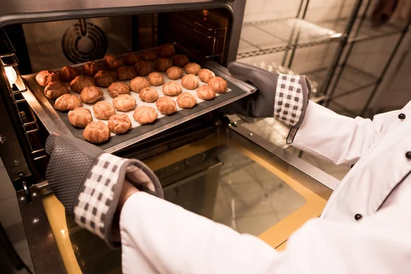 Partial view of confectioner taking eclairs on baking pan out of oven — Stock Photo